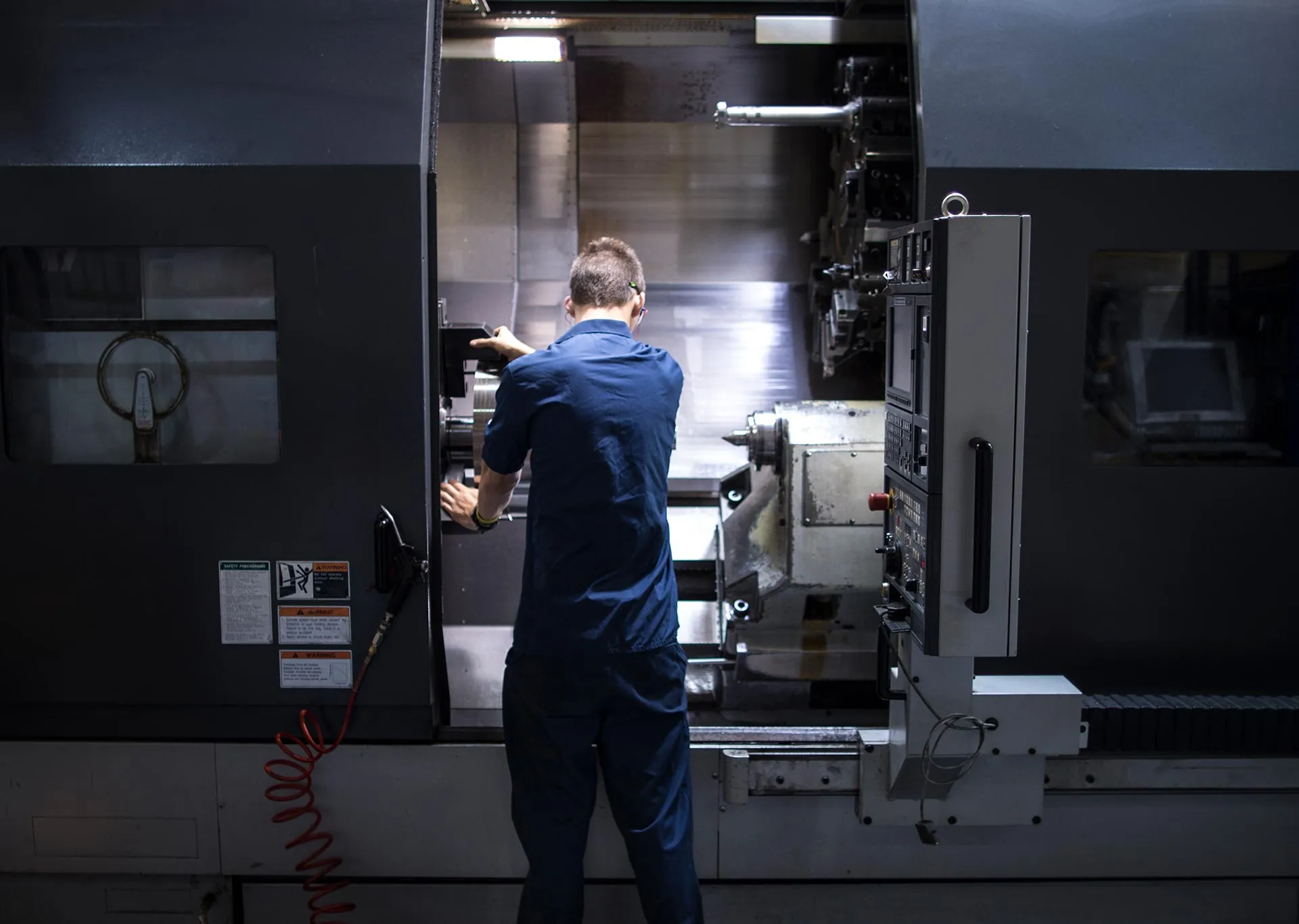 A machinist in navy coveralls loading a part into a CNC lathe, the machine's control panel lit beside him on a dark shop floor.