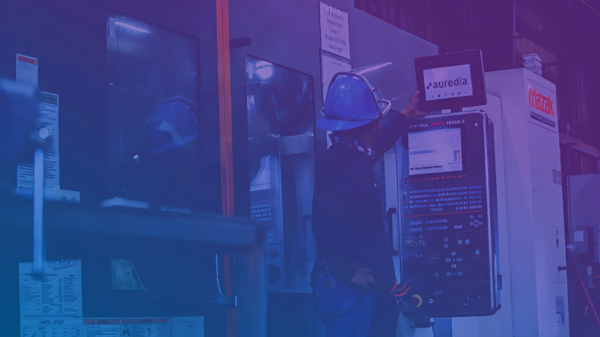 A machinist in a hard hat at the control panel of a Mazak CNC machine, holding a tablet running Auredia.